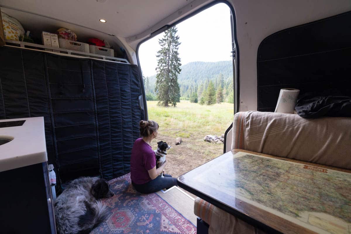 Kristen Bor sitting in the entryway of a Sprinter van witht he sliding door open and a dog in her lap while staring out at the view of trees