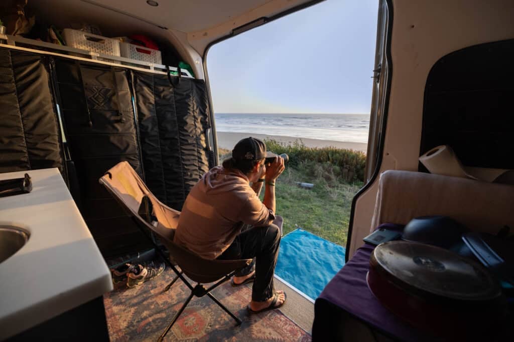 man sitting inside a sprinter van with binoculars parked on a cliff above the ocean at sunset in olympic national park