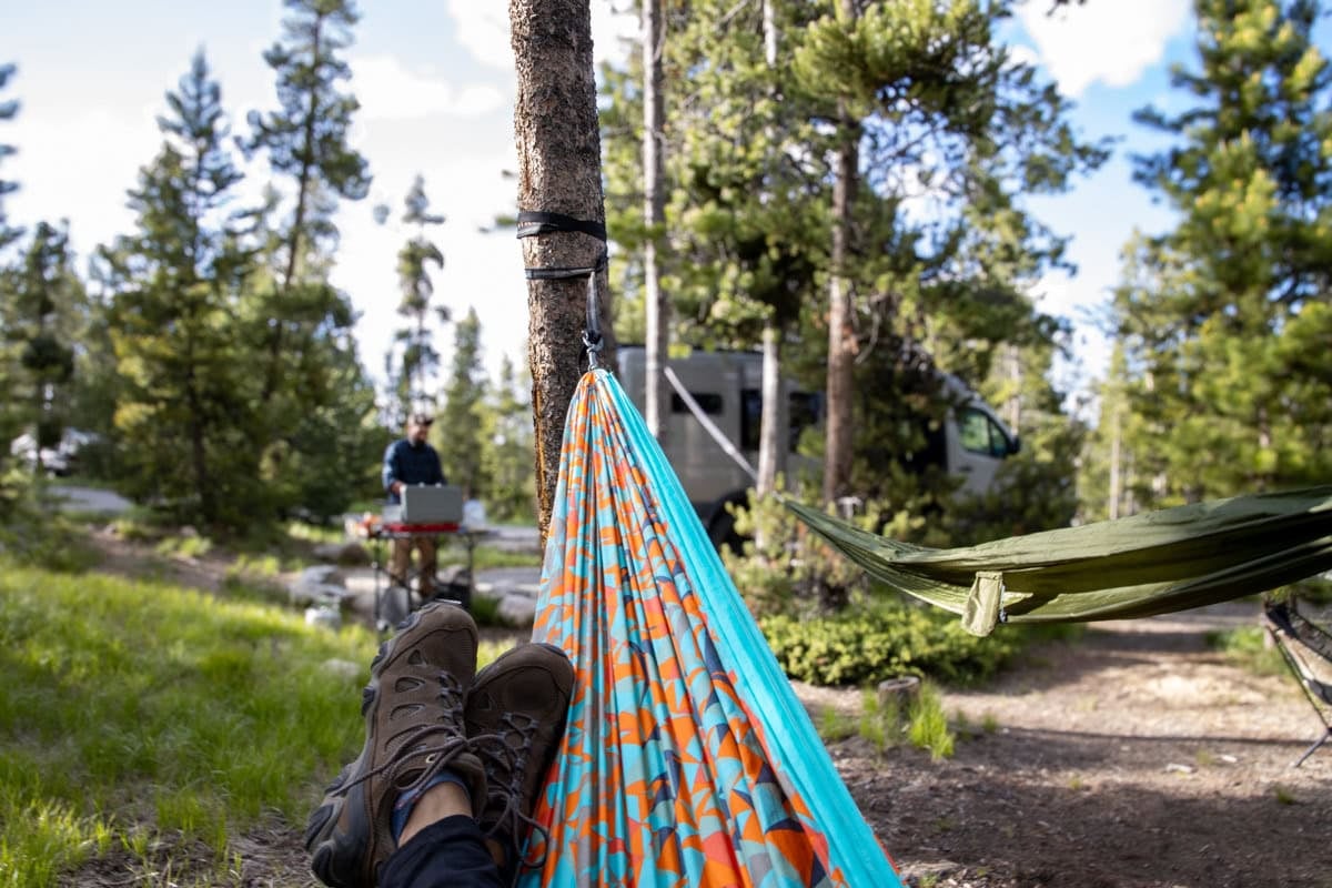 feet in a hammock with a man cooking on a camp stove and a sprinter van in the background