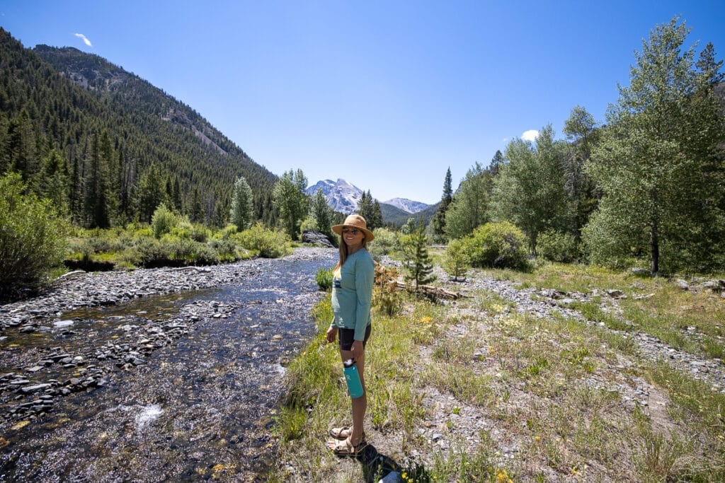 Kristen Bor standing next to a creek surrounding by forested mountains holding a Hydroflask water bottle