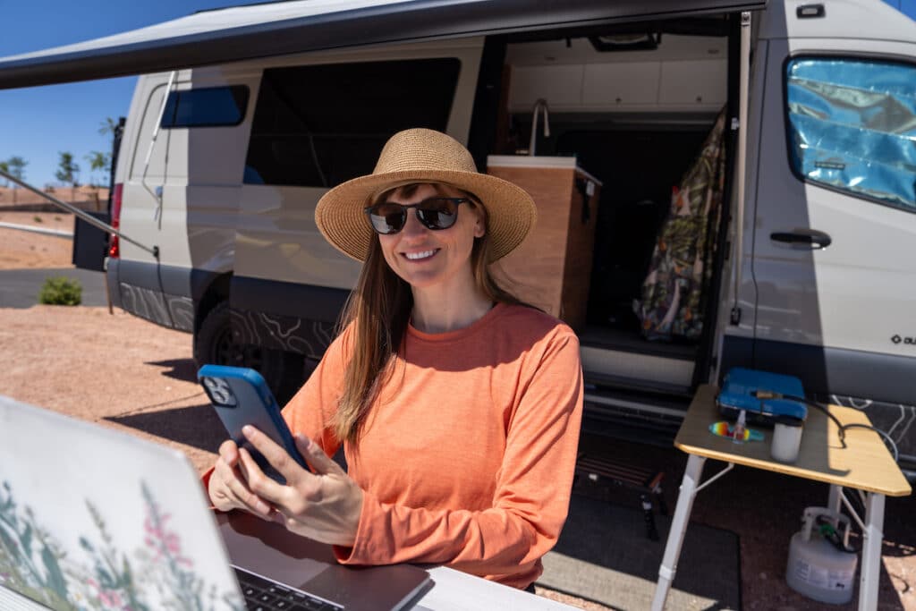 Kristen Bor with a laptop and her phone in her hand sitting at a picnic table that is next to a sprinter van