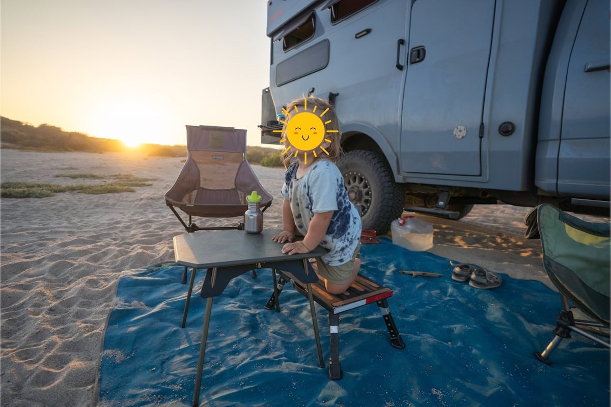Boy with face blocked out at a campsite on the beach next to a Rossmonster Baja Truck camper. Boy is sitting at NEMO moonlander table