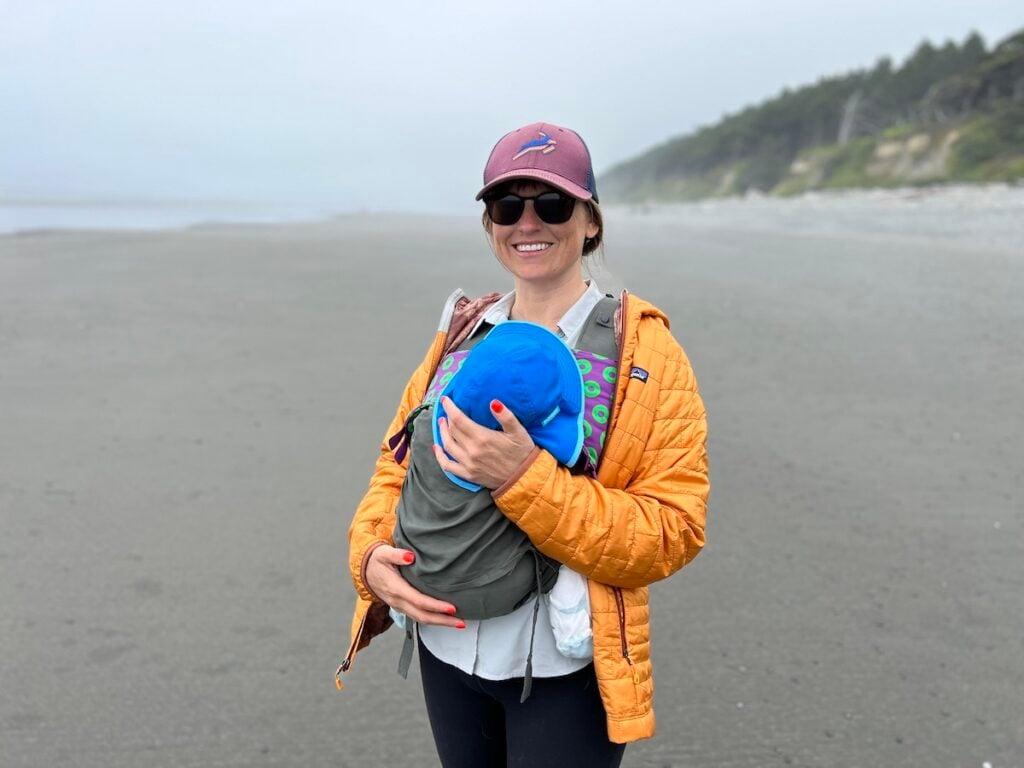 Bearfoot Theory founder Kristen Bor on a foggy beach in Olympic National Park wearing a gold Patagonia Nano puff and holding her baby in a carrier